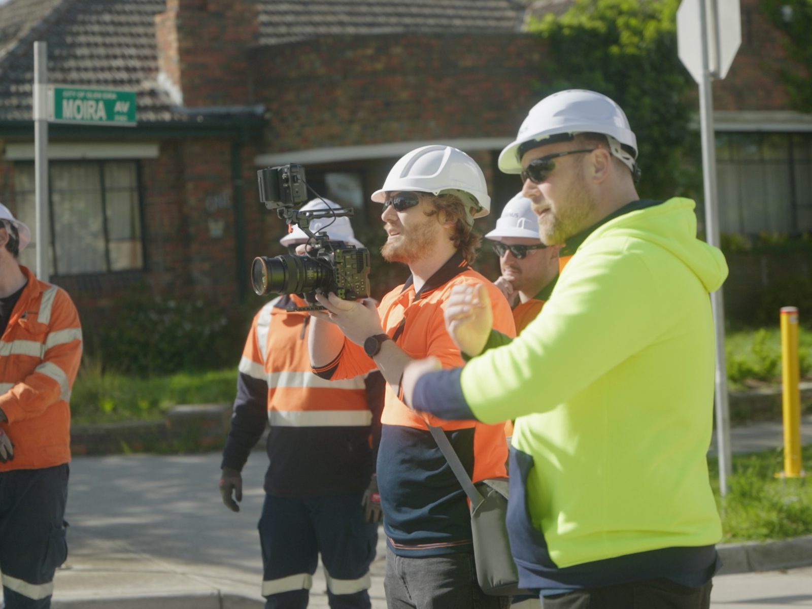 A Lift camera operator filming a crew in hard hats and hi-vis on a suburban street