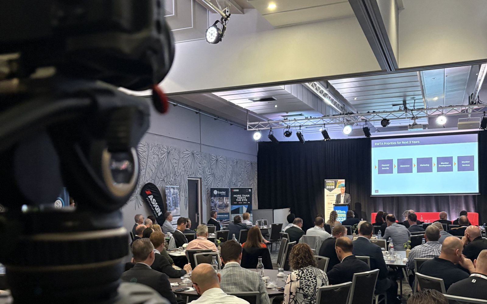 A Lift camera on a tripod at the back of a conference room, filming a speaker and a full audience