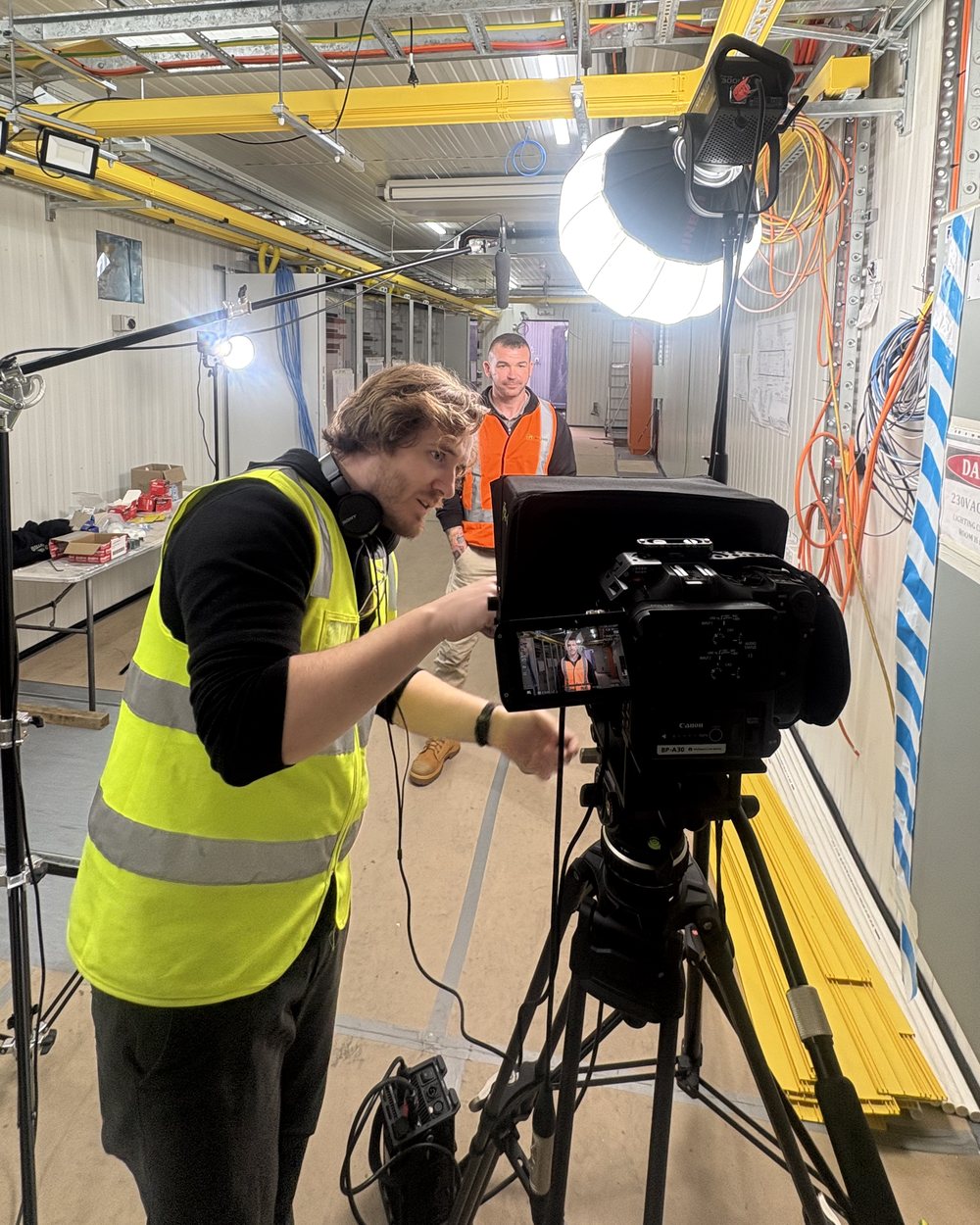 A Lift camera operator setting up an interview with a worker in hi-vis inside an electrical switchroom