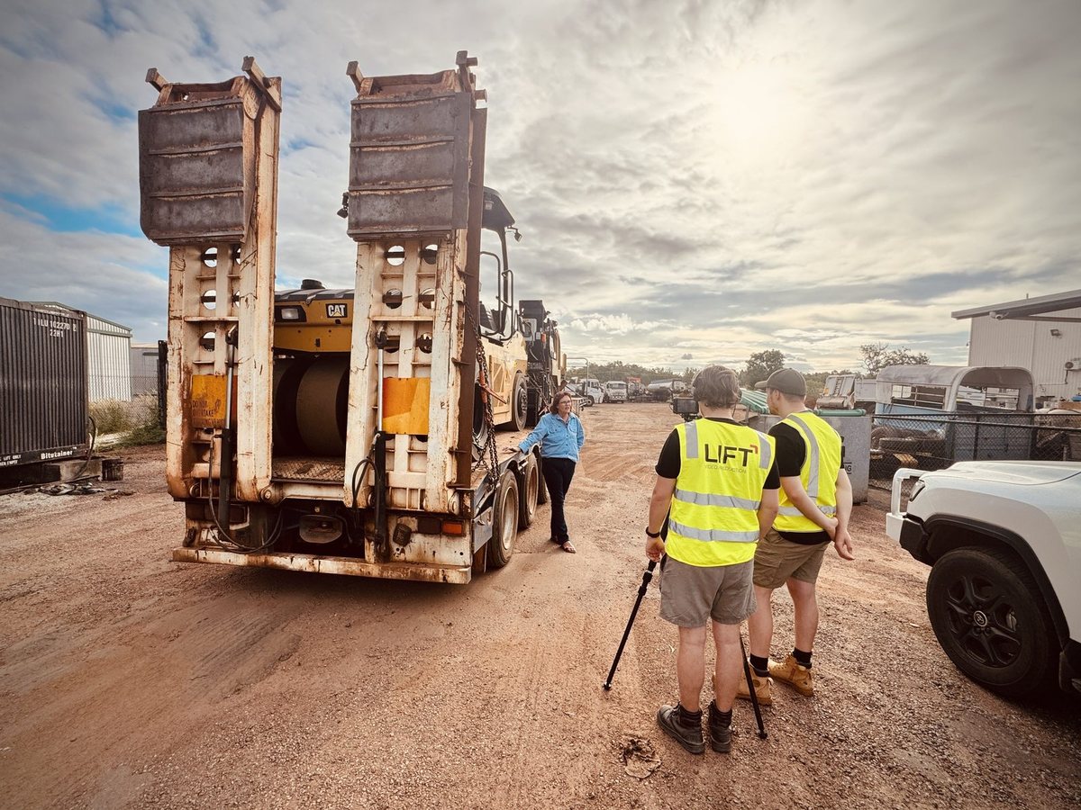 Two of the Lift crew in hi-vis vests on red dirt behind a low loader, filming for FJ Bitumen