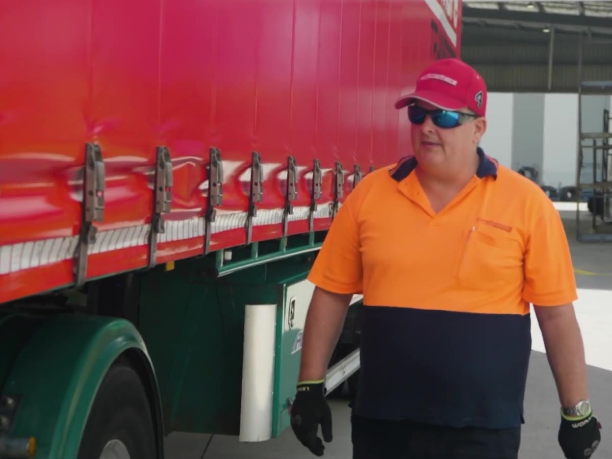 A truck driver in orange hi-vis doing a walk-around check beside a curtain-sider
