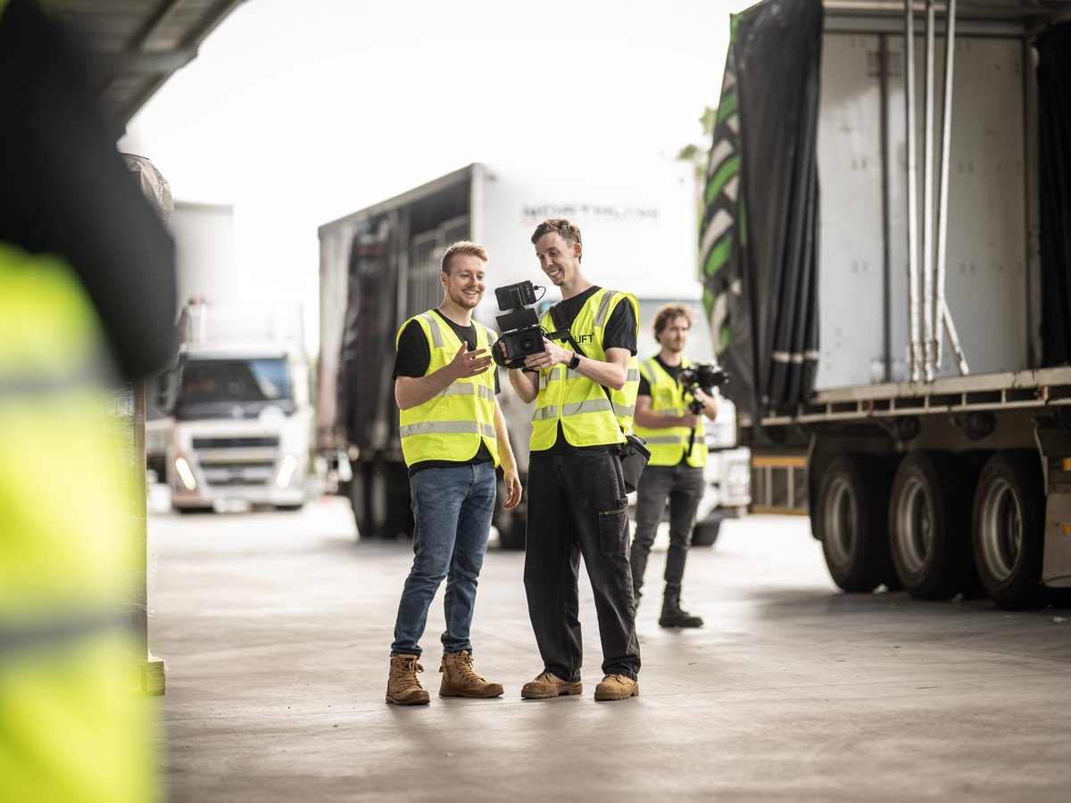 Three of the Lift crew with a cinema camera in a transport yard, trailers and a prime mover behind them